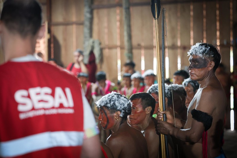 Um grupo de homens indígenas com pintura facial está reunido em círculo. Ao lado deles, à esquerda da foto, um homem de costas veste um colete vermelho com a sigla SESAI, da Secretaria de Saúde Indígena.
